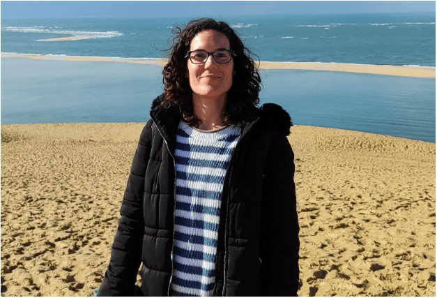 Foto de Laura Moreno Huete en un paisaje de playa invernal. Laura tiene el cabello rizado a la altura de los hombros, usa gafas y lleva una camiseta a rayas horizontales azules y blancas y una chaqueta negra abrigada. Mira de frente a la cámara y sonríe.