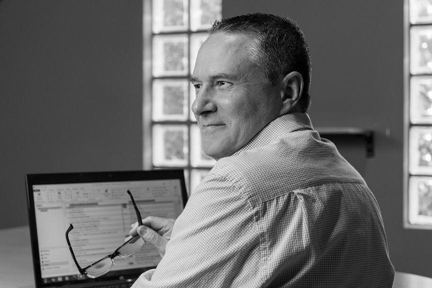 Foto en blanco y negro de Willy Martinez, un hombre de mediana edad. Se lo ve sentado de espaldas frente a una computadora portátil. Está mirando hacia la izquierda, por lo caual podemos ver tres cuartos de perfil de su rostro. Sonríe y sostiene un par de anteojos.