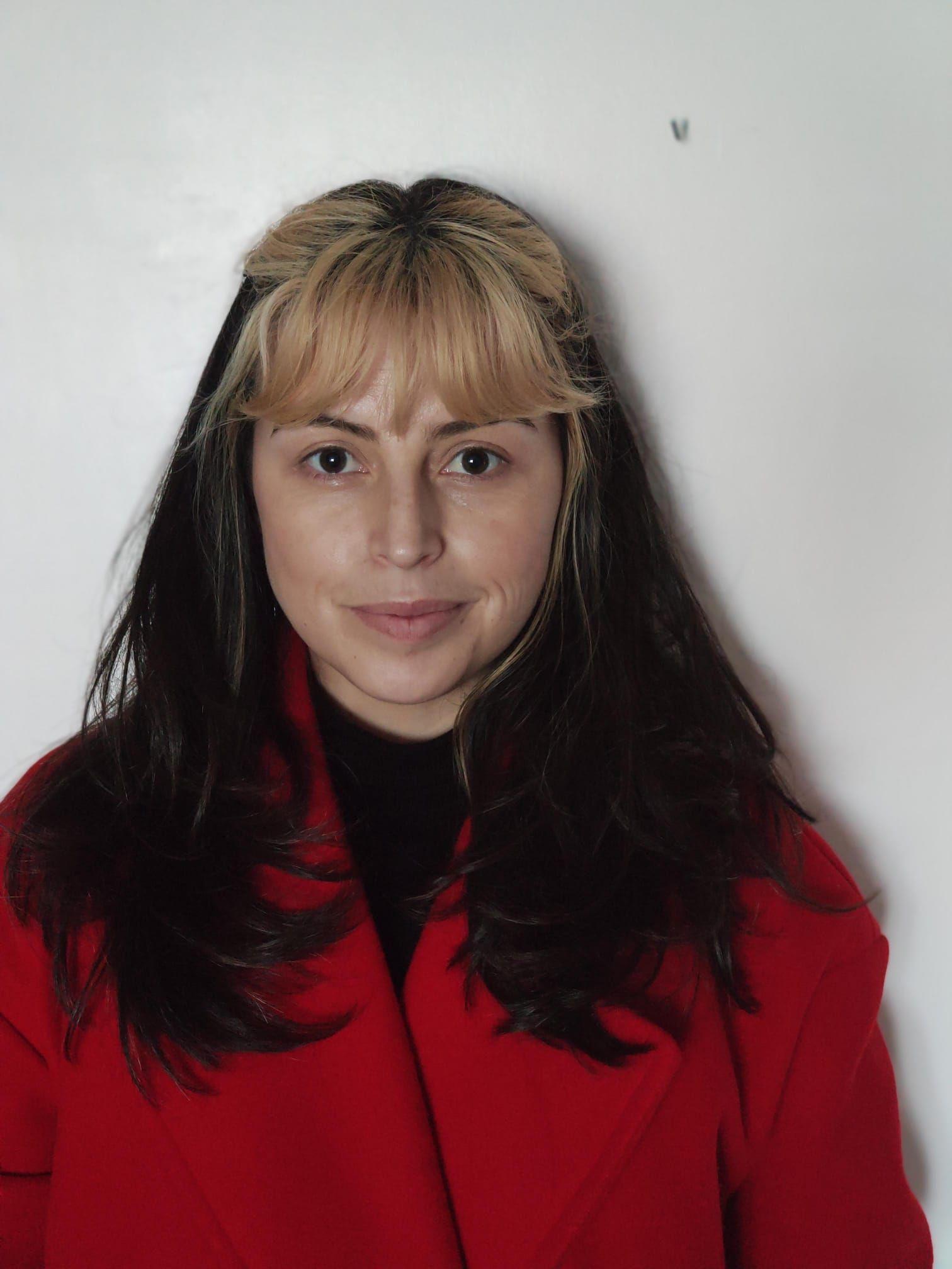 Plano medio de Laura Victoria Lesmes Velasco. Tiene el cabello largo, liso y castaño oscuro, salvo el flequillo que está decolorado. Es una mujer de 35 años. Lleva una camiseta negra debajo de un saco rojo. Mira a la cámara, con una media sonrisa.