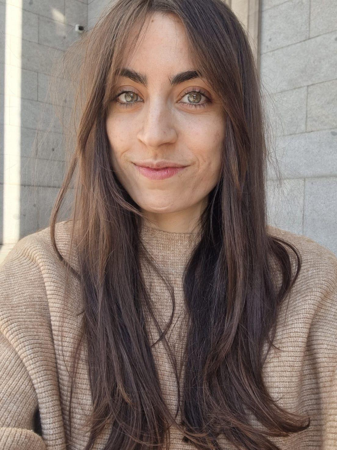 Foto de Cristina Joven. Cristina es una mujer de alrededor de 35 años. Tiene cabellos largos castaños y lacios; tez clara y ojos verdes. En la foto mira de frente a la cámara y sonríe. Lleva puesto un pullover en tono marrón claro.