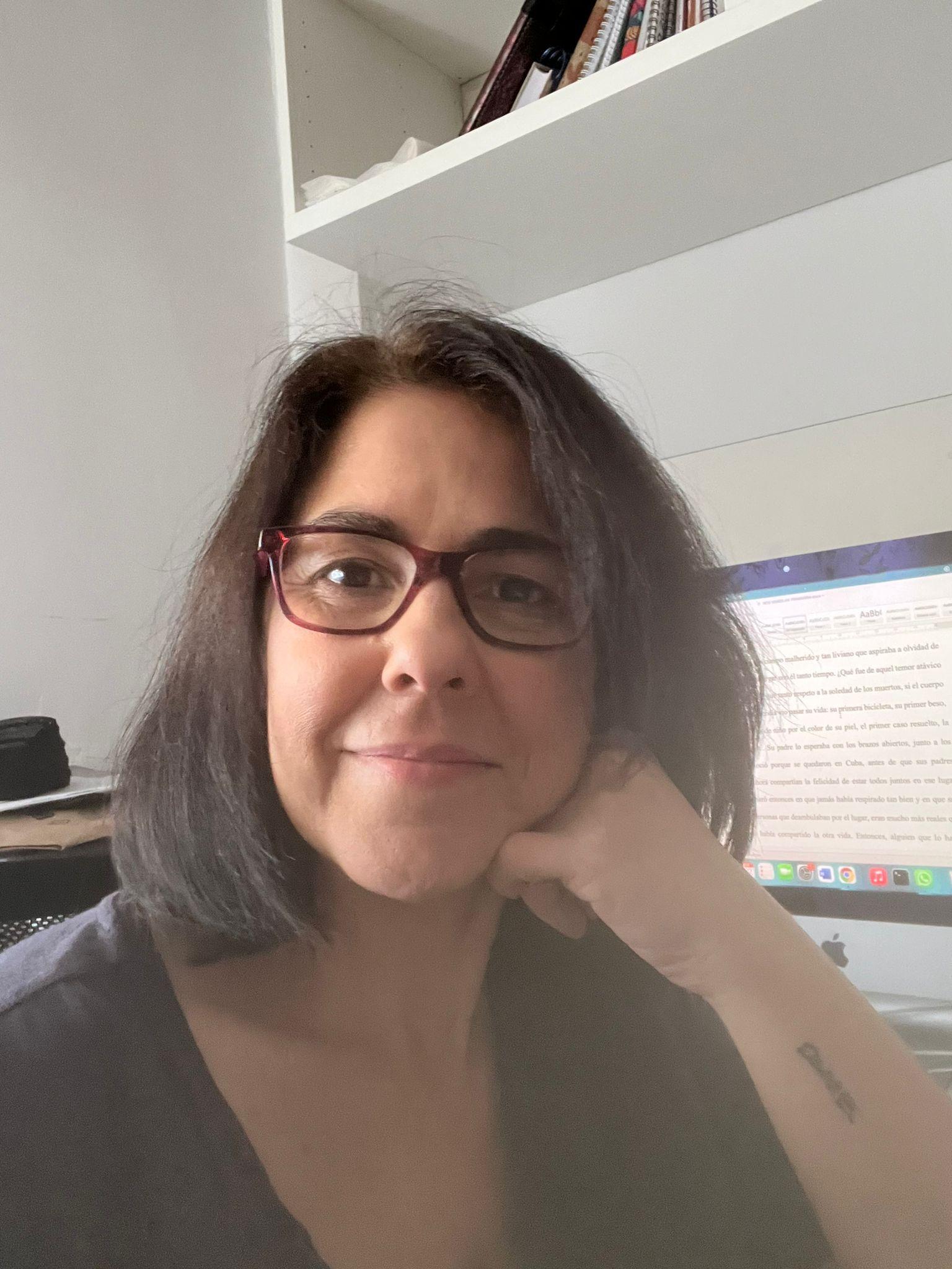 Primer plano de Gema Alcalá Recuero. Gema tiene tez clara, usa gafas y tiene el cabello liso, castaño y corto a la altura de los hombros. Mira de frente a la cámara, tiene la barbilla apoyada sobre la mano izquerda y sonríe ligeramente.